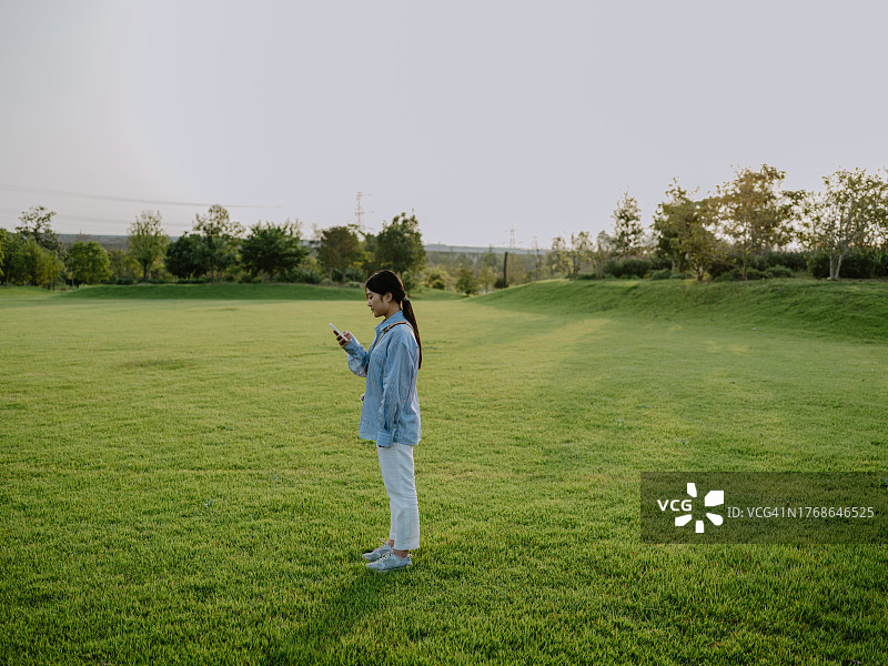 年轻的亚洲女子站在自然公园的草地上玩智能手机图片素材