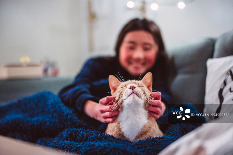 快乐的十几岁的女孩抓她的姜色虎斑猫的脸，晚上一起坐在沙发上毯子。爱护宠物，享受与宠物猫的亲密时光。图片素材