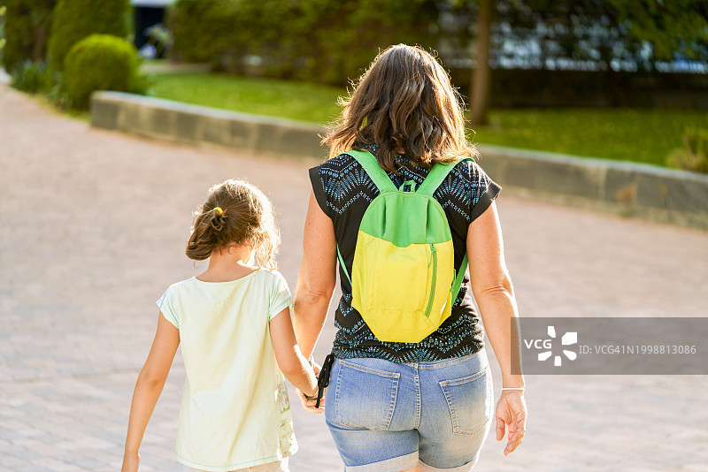 母亲和女儿穿着便服，背着背包，在公园里手牵手散步(后视图)。图片素材