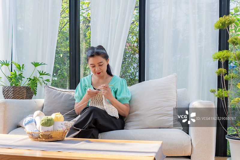 一位中国退休老妇人穿着绿色休闲服装坐在舒适的沙发上，在家里的客厅用钩针编织一条长长的米色围巾。篮子里彩色毛纱的线索。室内放松活动。周末的休闲活动。图片素材