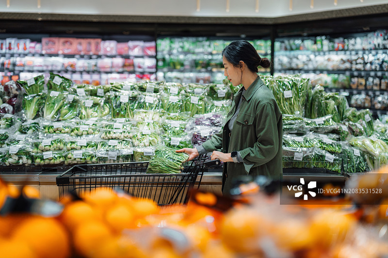 年轻的亚洲妇女在超市购买新鲜的有机水果和蔬菜。把一袋蔬菜放进购物车。食品杂货店购物。健康的饮食习惯。吃绿色可持续的饮食方式图片素材