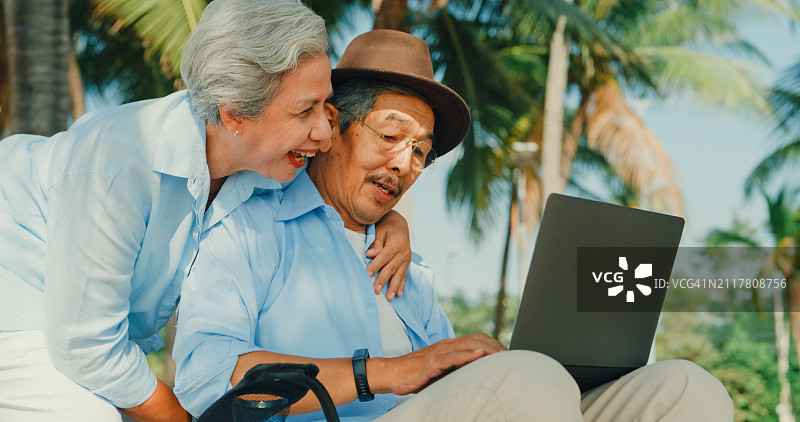 一位亚洲老人坐在沙滩椅上玩笔记本电脑，和妻子一起欣赏海景。一对幸福的老年夫妇享受退休后的旅行生活方式。图片素材