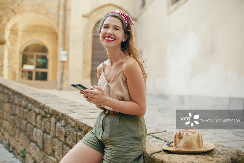 可爱的年轻女游客坐在石架上，拿着手机，对着镜头笑得很开心图片素材