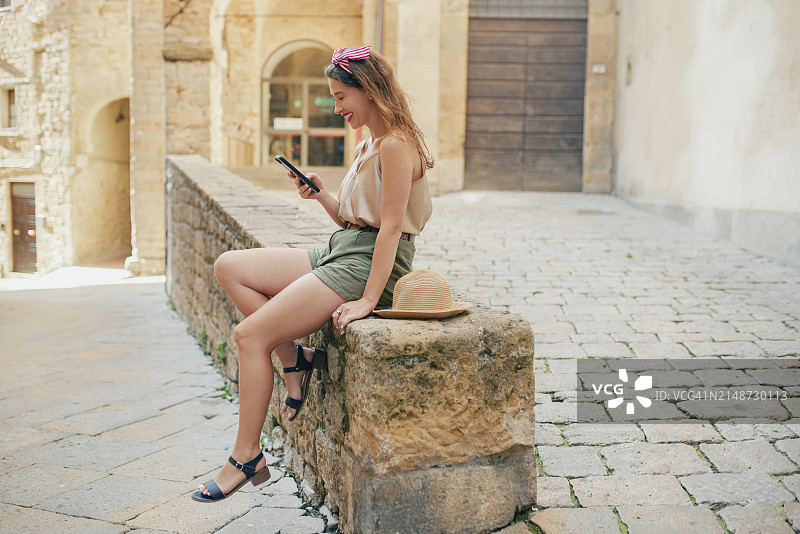 中世纪村庄里，年轻女游客坐在石壁上使用手机的侧视图图片素材