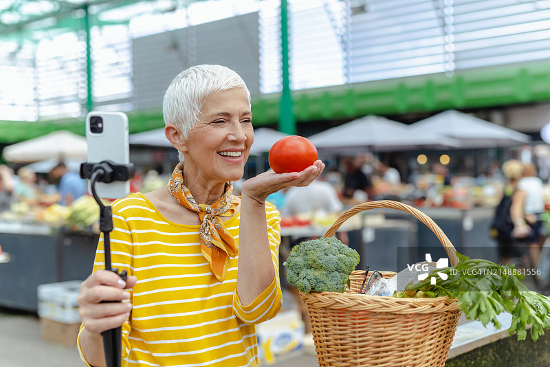 一位穿着讲究的白人老年妇女在市场上拿着自拍杆，用手机自拍图片素材