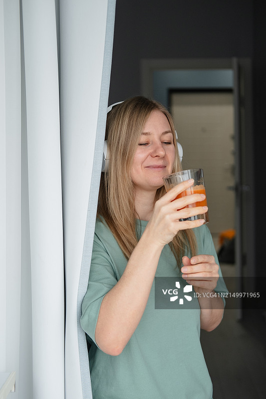 一个戴着无线耳机的快乐女人站在阳台上，手里拿着橙汁图片素材