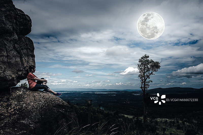 坐在圆石上的女人，天空阴云密布，月圆满满。图片素材