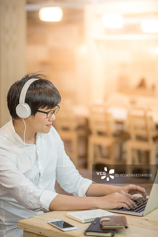 亚洲快乐的年轻人戴着耳机听音乐，用笔记本电脑工作图片素材