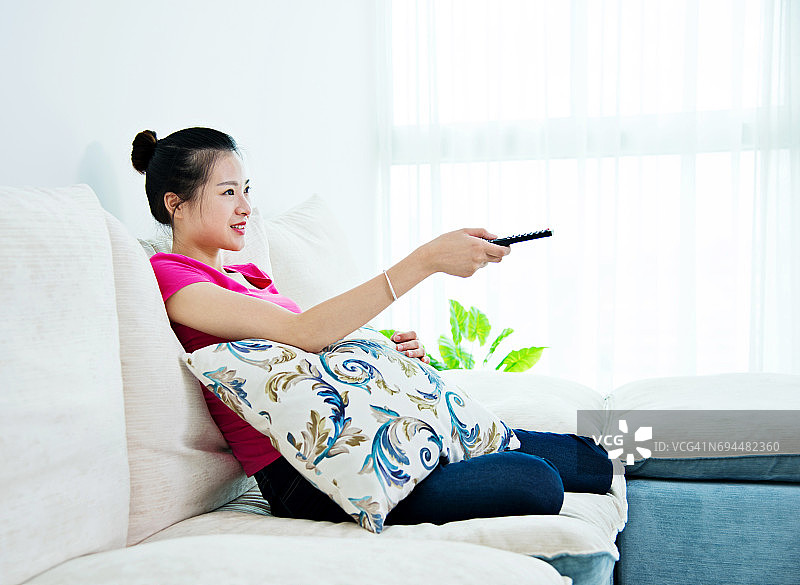 年轻的亚洲女人用遥控器在沙发上看电视图片素材