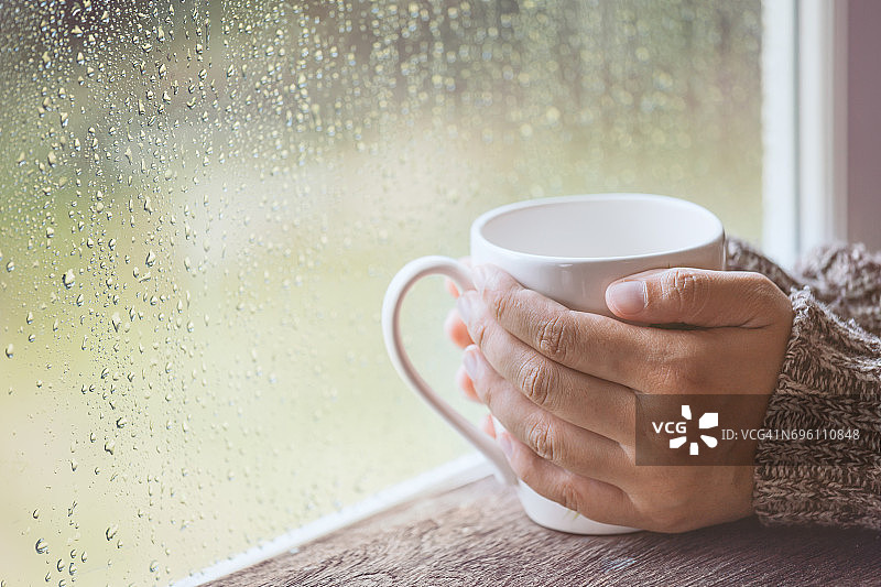 女人手拿着一杯咖啡或茶在雨天的窗户背景图片素材