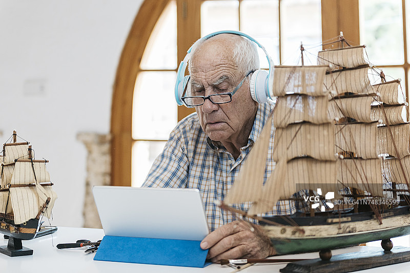 戴着耳机的老人在桌子上用平板电脑制作船模型图片素材