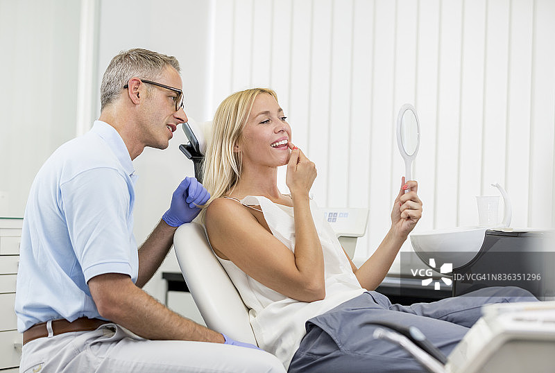 牙医的办公室美学美容牙科-短头发花白的头发的中年人牙医咨询与长金发女病人的椅子上,医生描述了漂白增白的过程图片素材