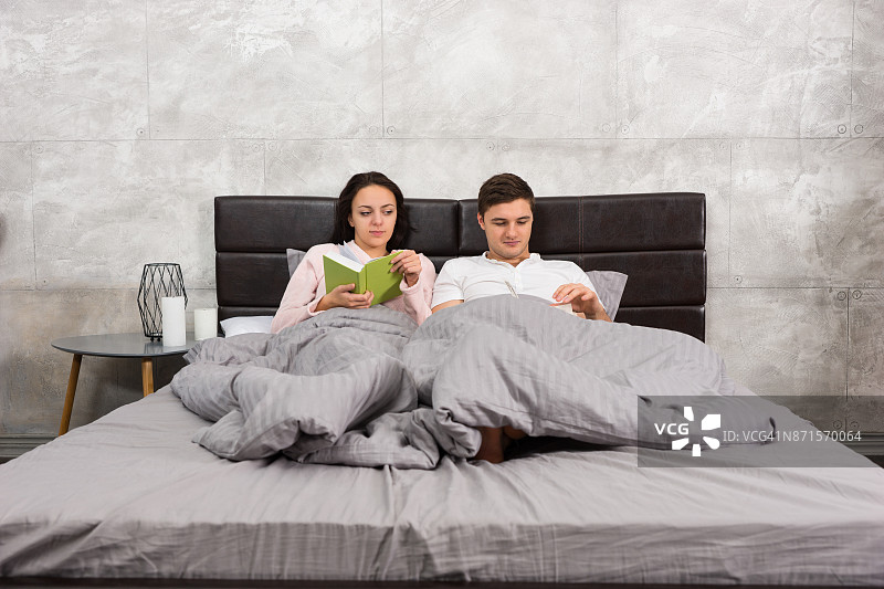 一对年轻夫妇穿着睡衣躺在床上看书图片素材
