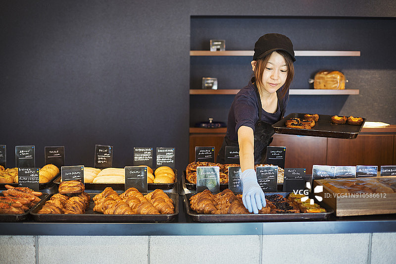 一位在面包店工作的妇女，正在把刚烤好的牛角面包和蛋糕放在柜台上的大托盘上。图片素材
