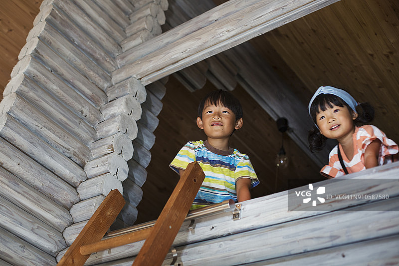 低角度观看男孩和年轻女孩的黑发看着窗外，梯子靠在木墙。图片素材