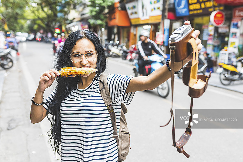 越南，河内，一名年轻女子在街上吃着玉米棒，用老式相机自拍图片素材