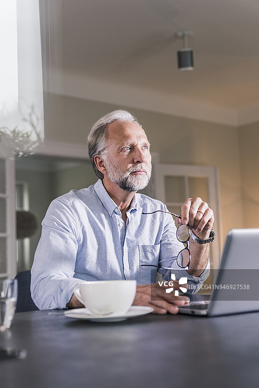 一幅沉思的成年男子的肖像，他坐在桌边，用笔记本电脑看着远方图片素材