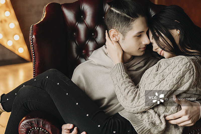 年轻可爱的夫妇面对面坐在皮革椅子上的肖像，而男人是抱着她的女朋友在他的胳膊。图片素材