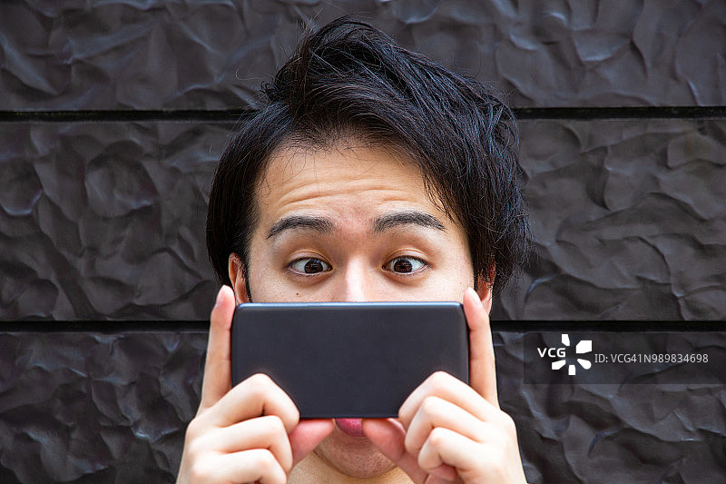 年轻的亚洲人看着智能手机，脸上带着惊讶的表情。图片素材