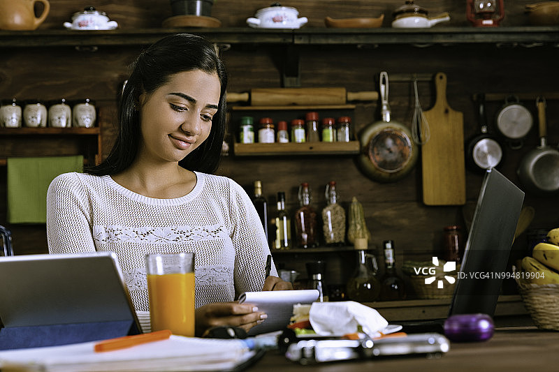 家庭办公室工作。西班牙裔年轻女子在厨房用笔记本做笔记图片素材