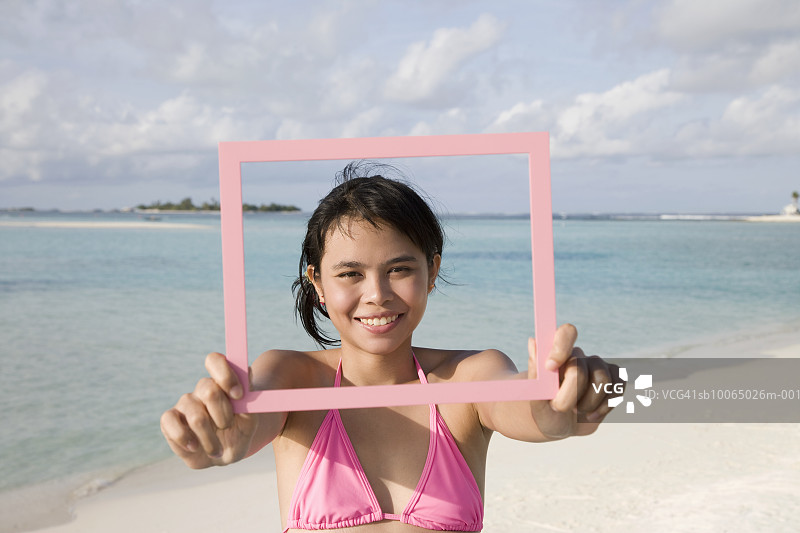 年轻女子在海滩上，拿着相框遮住脸，肖像图片素材