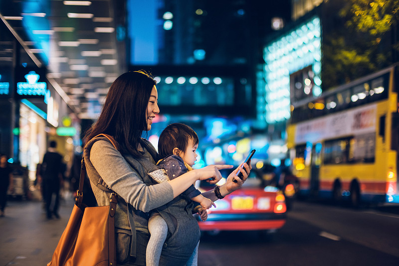 年轻的亚洲母亲带着女儿在市中心的街道上用智能手机应用安排出租车，背景是模糊的交通场景图片下载