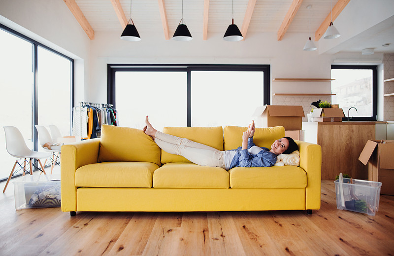 一名年轻女子拿着智能手机躺在新家现代客厅的黄色沙发上，旁边是移动的箱子。图片下载