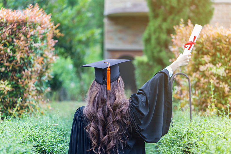 后视图的女人在毕业礼服持有证书而站在植物图片素材