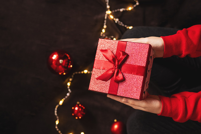 美丽的圣诞礼物带着一个红色的蝴蝶结，在一个年轻女孩的手中。红毛衣，圣诞球和彩灯。图片下载