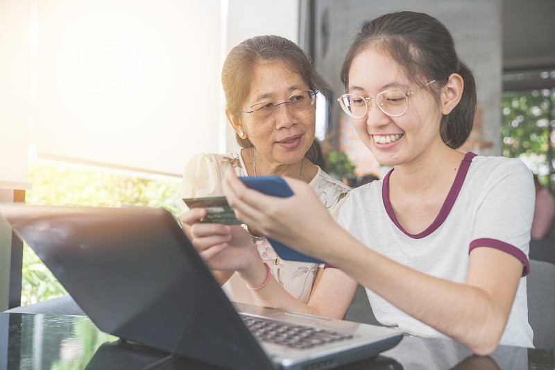 亚洲家庭年轻的青少年和母亲度假放松使用数字平板电脑和智能手机在家里做作业，放松与网络游戏和短信在社交网络使用信用卡图片下载