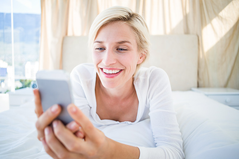 微笑的金发女人躺在床上，用她的手机在卧室发短信图片下载