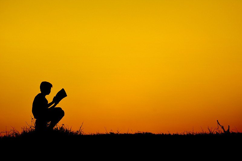 天空日落时，一个正在看书的男孩的剪影图片素材
