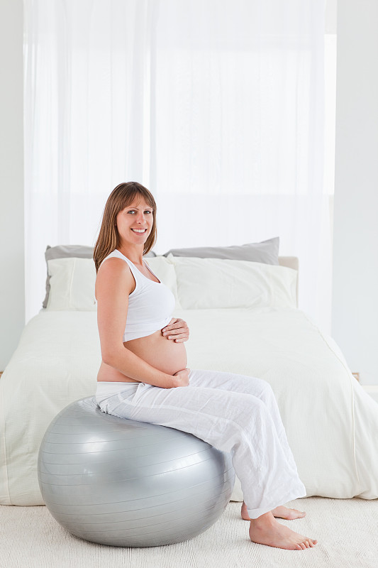 美丽的孕妇坐在卧室的健身球上抚摸着自己的腹部图片下载