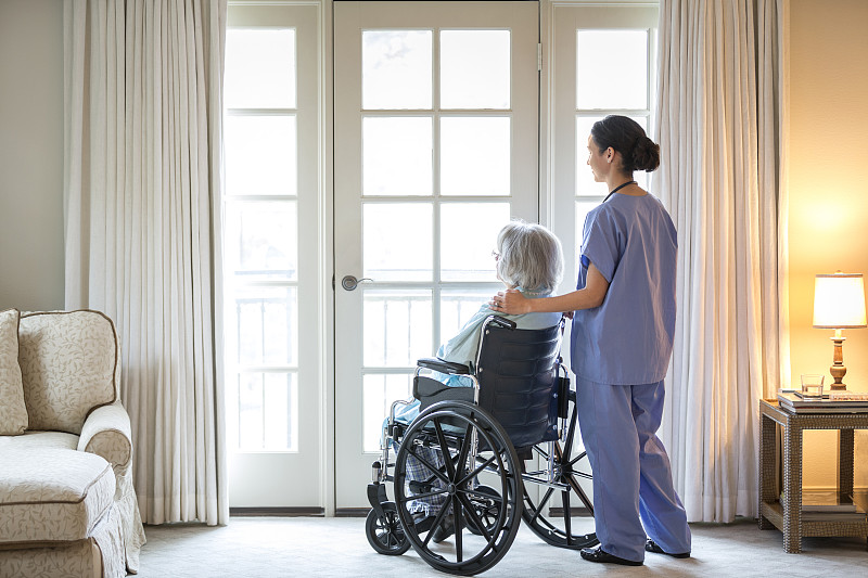 护士和年长病人坐在轮椅上，靠窗站着图片下载