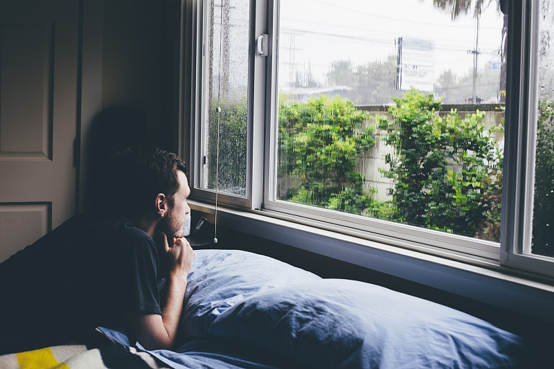 男人躺在床上，透过窗户望着雨图片下载