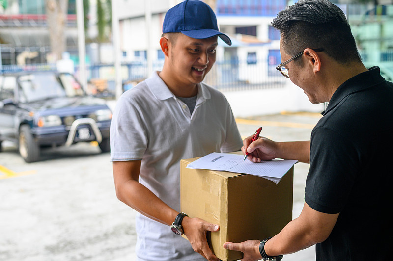 亚洲快递员微笑着拿着一个纸板箱给他的客户。图片下载