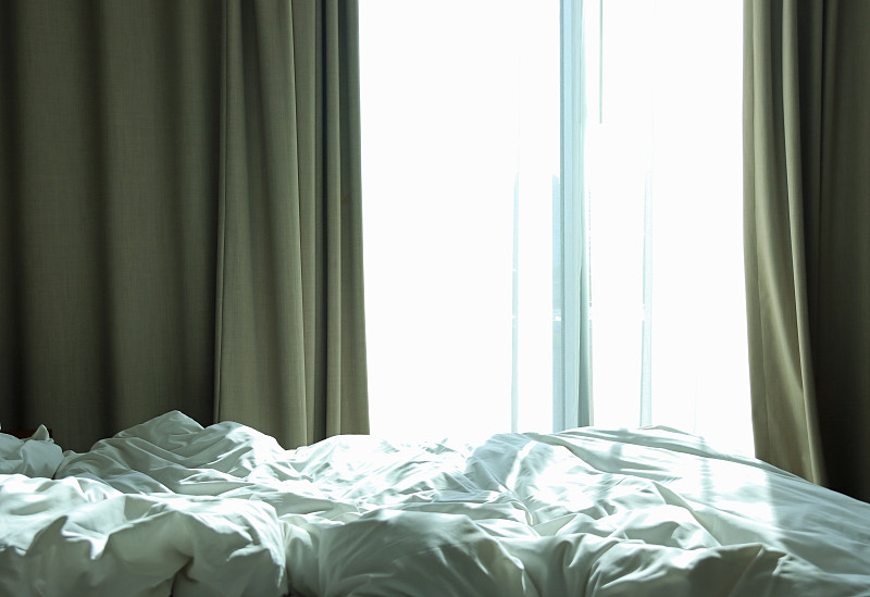 酒店的窗户拉上了窗帘，让明亮的晨光照进来，前景是凌乱的床图片下载
