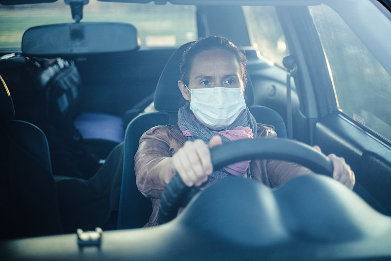 活跃的年轻女子开车去上班。戴着保护面罩开车。图片素材