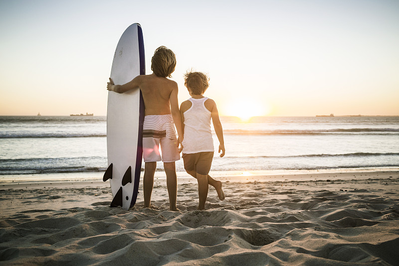 后视图的两个男孩与冲浪板站在海滩上日落图片素材
