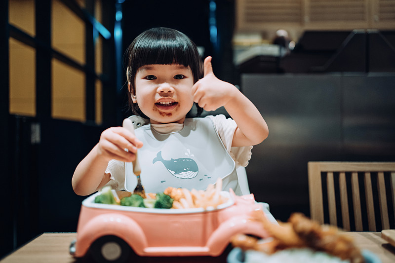 可爱的亚洲小女孩喜欢她的孩子套餐，在餐厅里用叉子吃自己的东西，同时用大拇指向上指图片素材