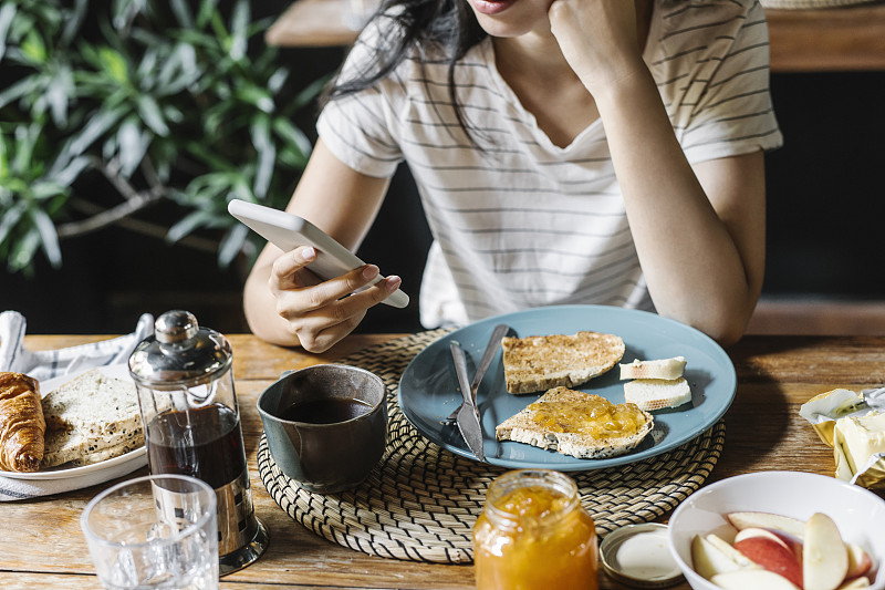 一个匿名女人使用智能手机在家里吃早餐的特写图片素材
