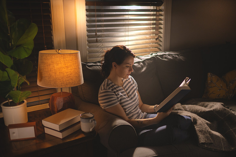 一个年轻女子坐在舒适的沙发上，在昏暗的光线下看书。图片素材