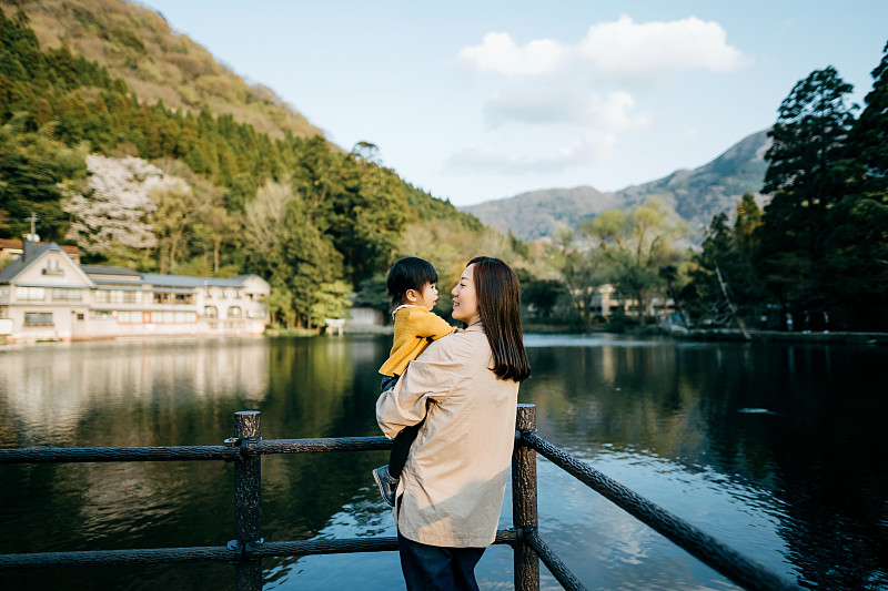 一位慈爱的年轻亚洲母亲抱着可爱的小女儿，在湖边观光，放松和享受美丽的自然风景。一起度过亲密的家庭时光图片下载