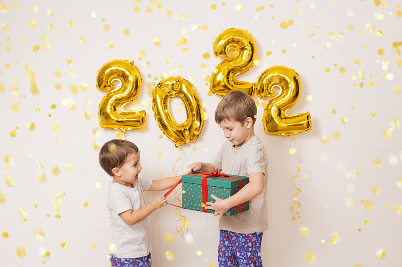 快乐微笑的孩子们拿着礼品盒，戴着红色的圣诞老人帽子，在白色的墙壁旁边，金色的气球享受着五彩纸屑。在家里庆祝2022年新年。圣诞节的概念图片下载