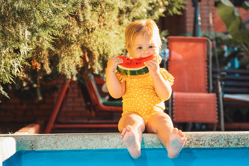 身穿黄色泳装的可爱白人小女孩坐在泳池边吃着一片西瓜。夏季图片下载