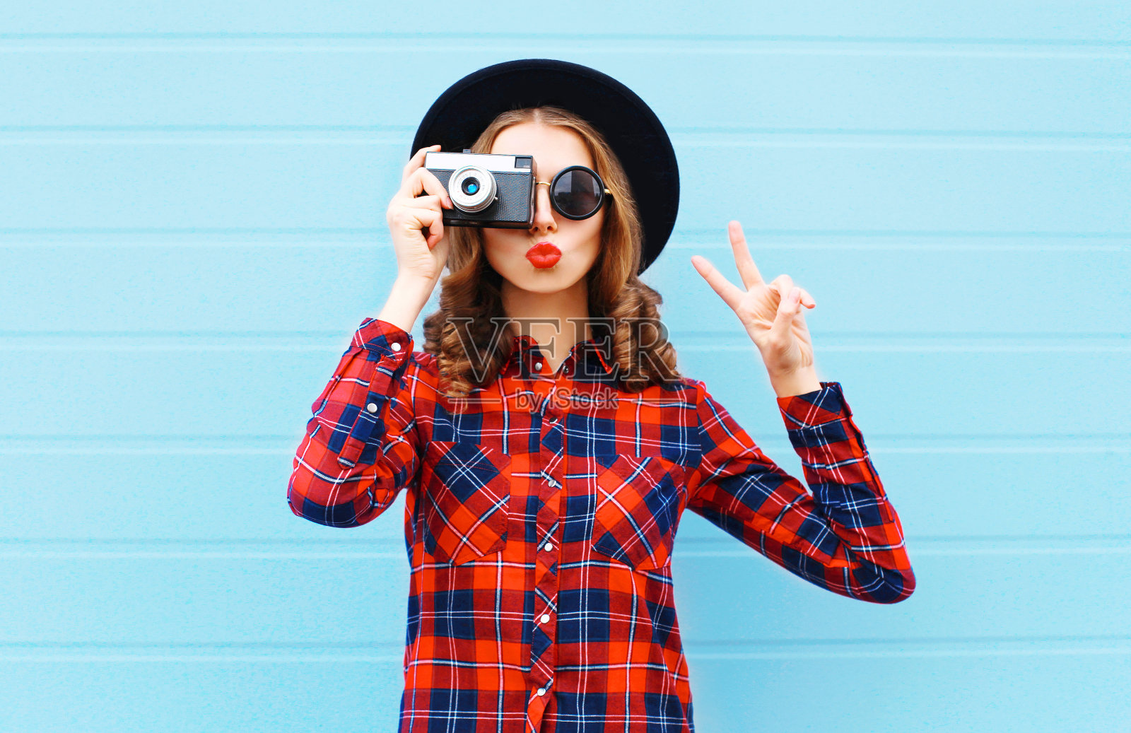 时尚漂亮的年轻女人吹红色的嘴唇与复古相机戴着黑色的帽子，红色格子衬衫在蓝色的背景照片摄影图片