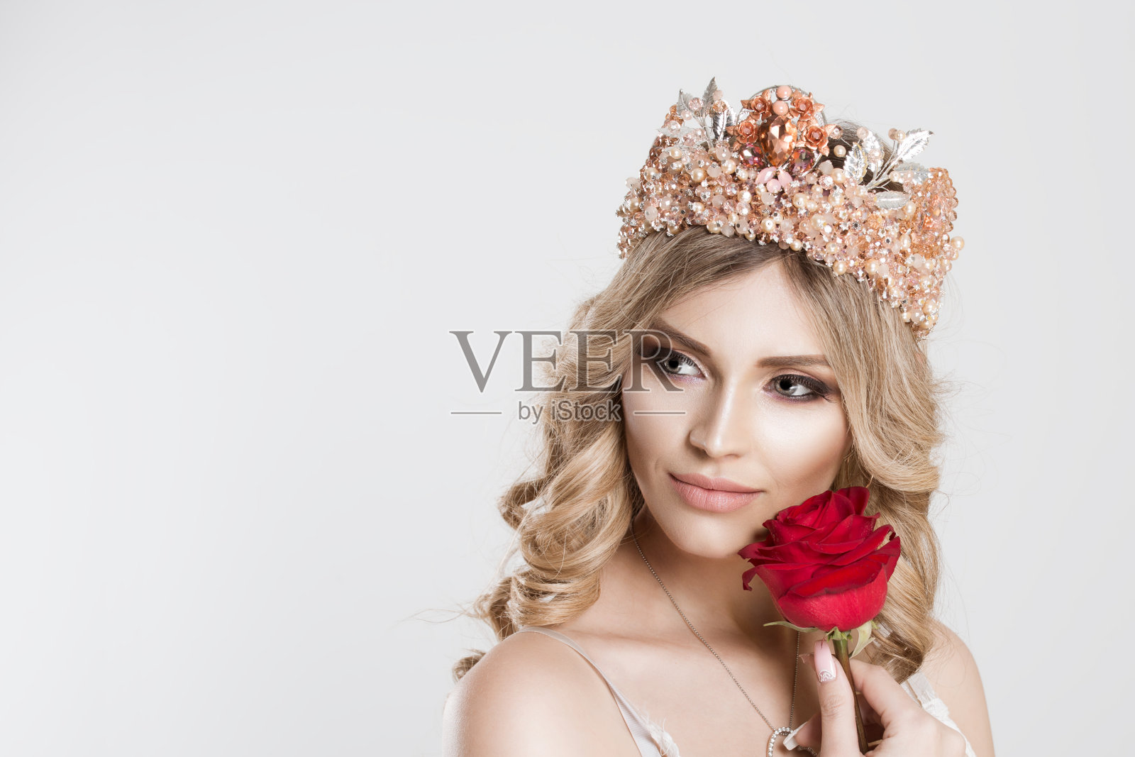美丽的皇后新娘与明亮的粉色皇冠举着红色玫瑰靠近她的脸颊看向一边，轻新娘妆孤立的灰白色背景墙照片摄影图片
