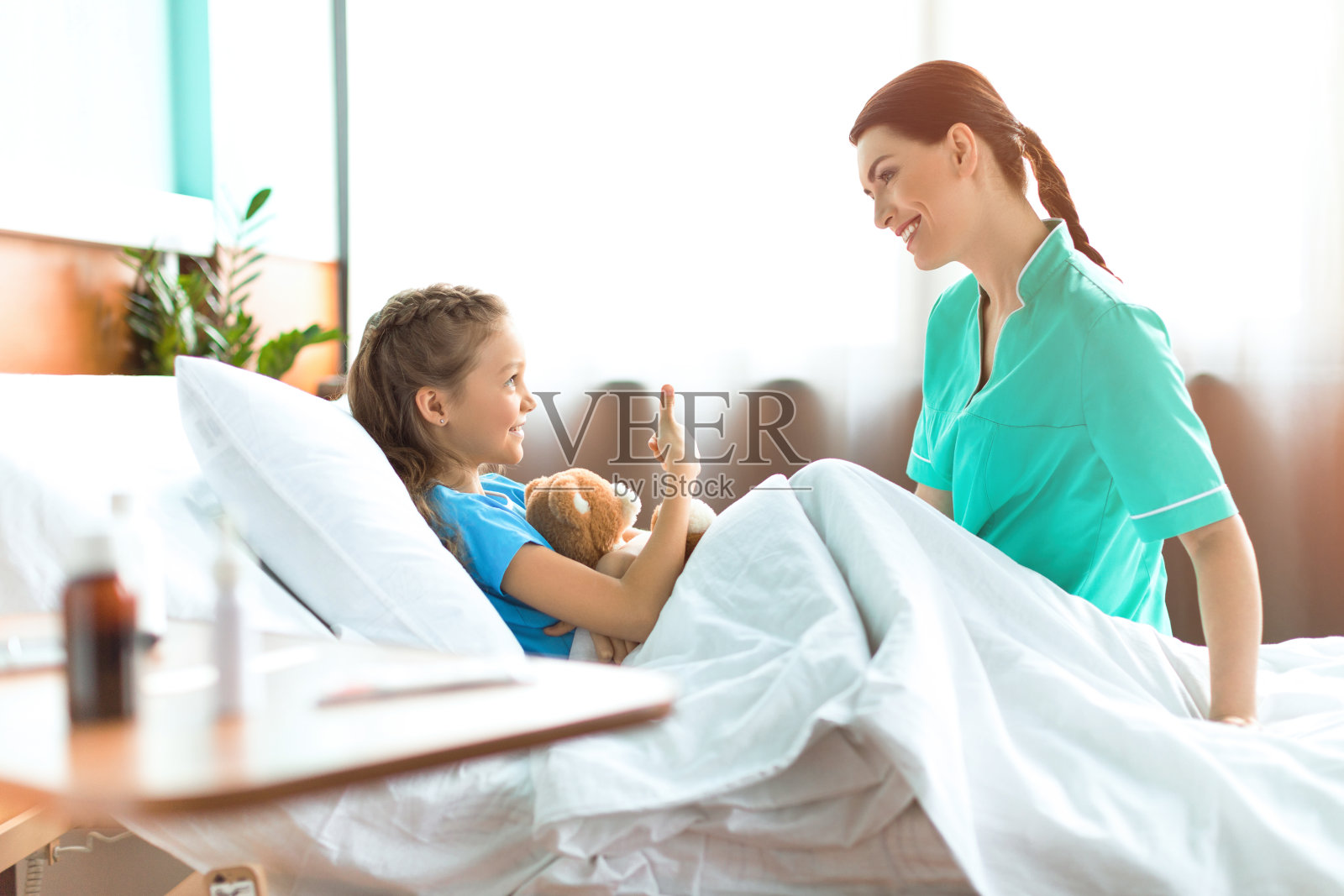 可爱的小女孩和泰迪熊躺在医院的床上，和微笑的护士交谈照片摄影图片