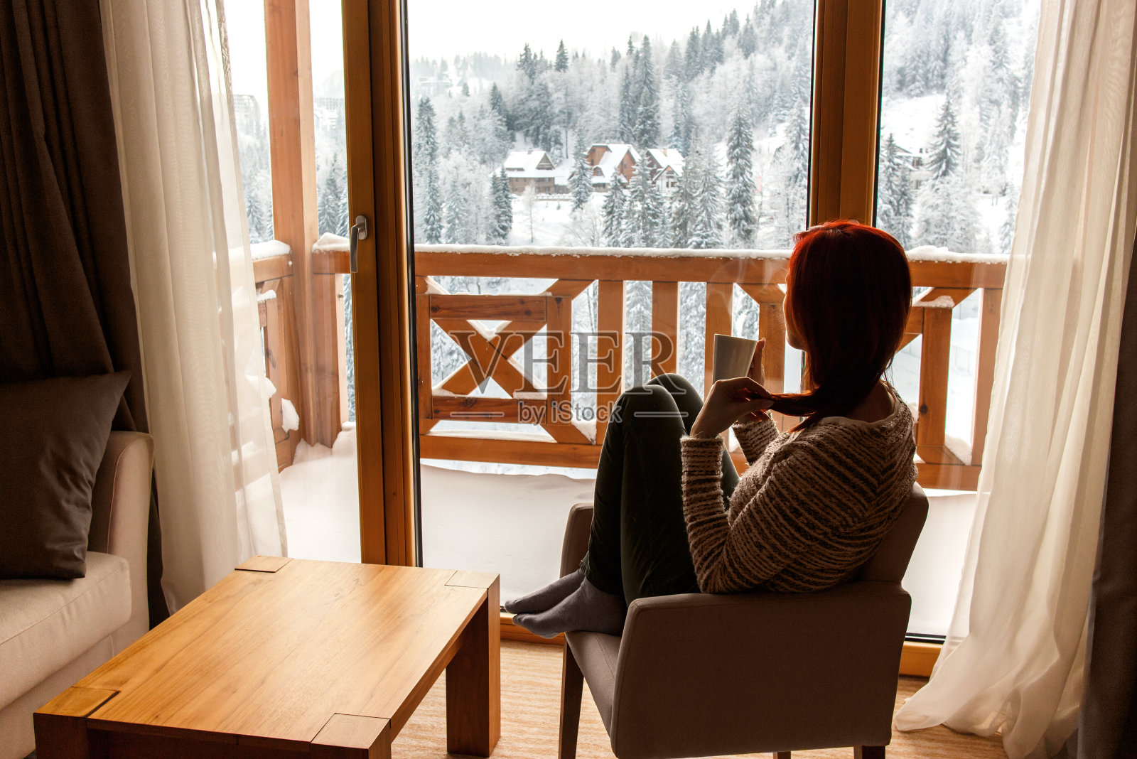 一个女人坐在舒适的椅子上，喝着咖啡，透过窗户看着白雪覆盖的山。照片摄影图片