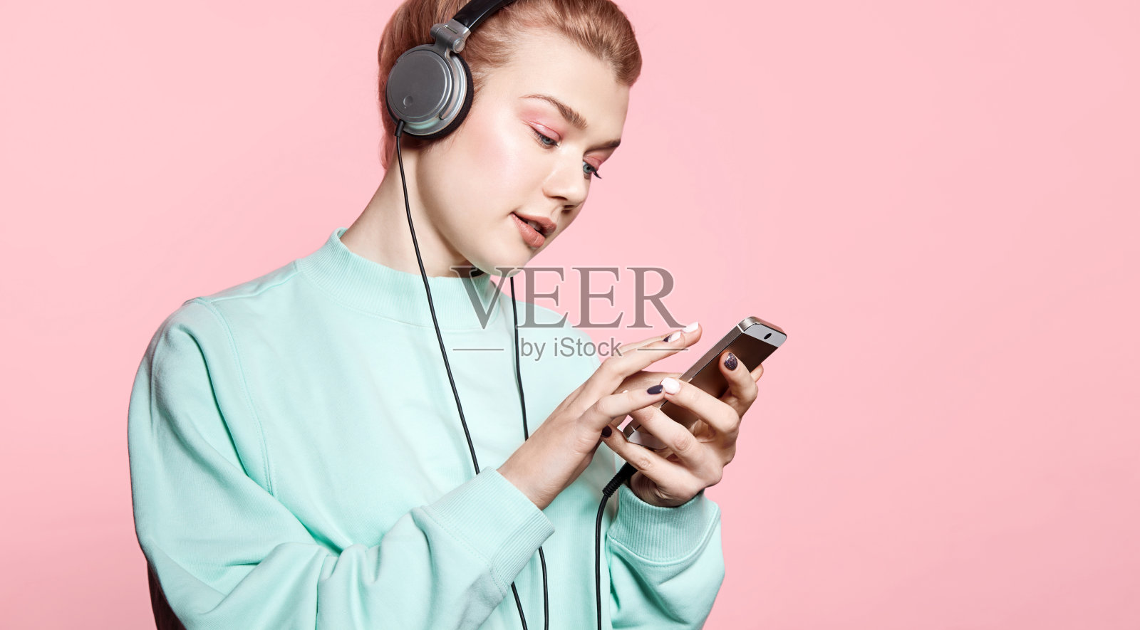 美丽的年轻女子戴着耳机听音乐，闭着眼睛站在粉红色的背景上，穿着蓝色的运动衫照片摄影图片